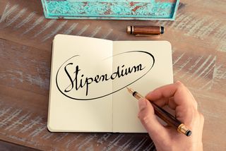 Image of a hand writing the German word for scholarship, "Stipendium", into a notebook with a fountain pen. The notebook is lying on a wooden table. Stock photo by user #224708 via Colourbox.