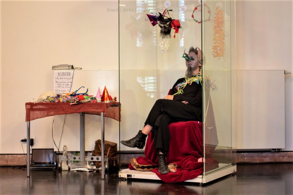 enlarge the image: Eine Studentin sitzt komisch verkleidet in einer Glasvitrine, daneben ein Tisch mit weiteren komischen Sachen